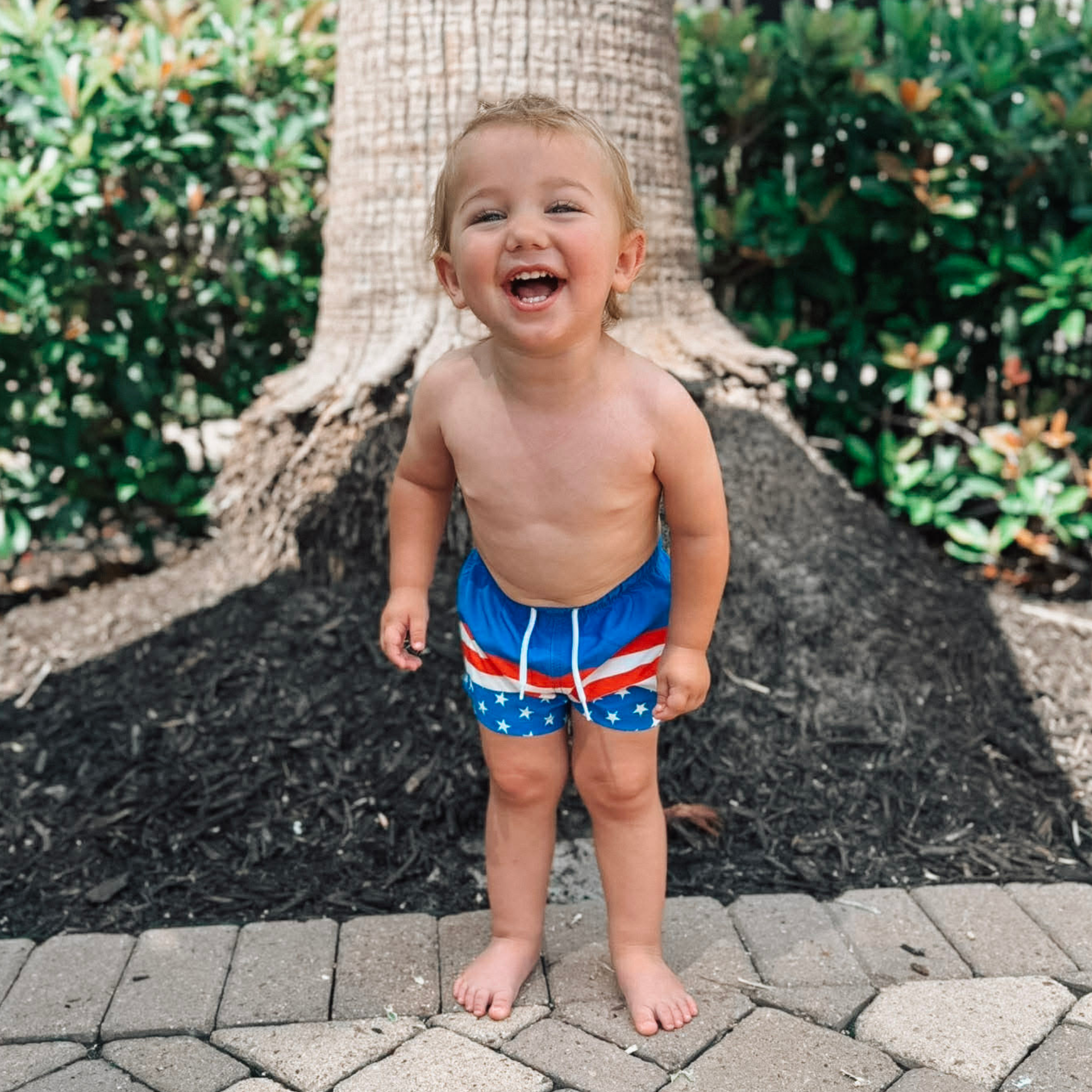Smiling toddler boy wearing Knuckleheads Retro USA flag swim shorts outdoors on a sunny summer day.