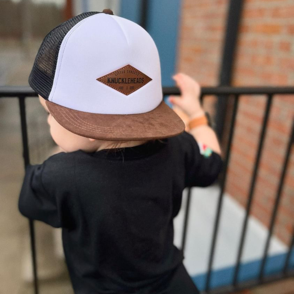 Image of White with Brown Bill Kids Trucker Hat with Troublemaker Patch: A cool and trendy trucker hat designed for kids. The hat features a stylish black crown and a brown bill, adorned with a striking Knuckleheads patch on the front. Elevate your child's style with this fashionable and comfortable accessory, perfect for any adventure or everyday wear. Crafted with care, this black with brown bill trucker hat with a Knuckleheads patch is a must-have addition to their wardrobe.