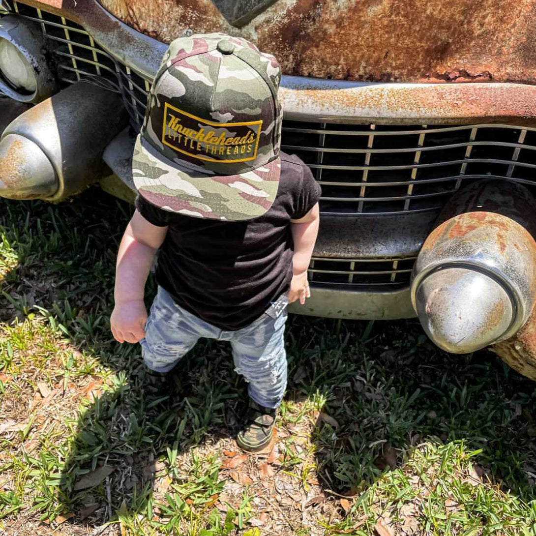 Image of Camo Kids Trucker Hat with Gold Rectangle Knuckleheads Patch: A rugged and stylish accessory designed for kids. In a classic camo pattern, it features a distinctive gold rectangle Knuckleheads patch on the front. Elevate your child's style with this fashionable hat, perfect for adding a touch of outdoor charm to their outfits.