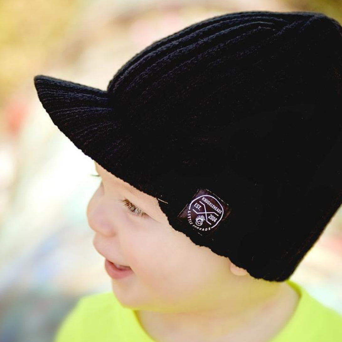 Young boy confidently sporting a black kids' beanie with a Knuckleheads tag and a stylish visor, showcasing a trendy and edgy fashion accessory.
