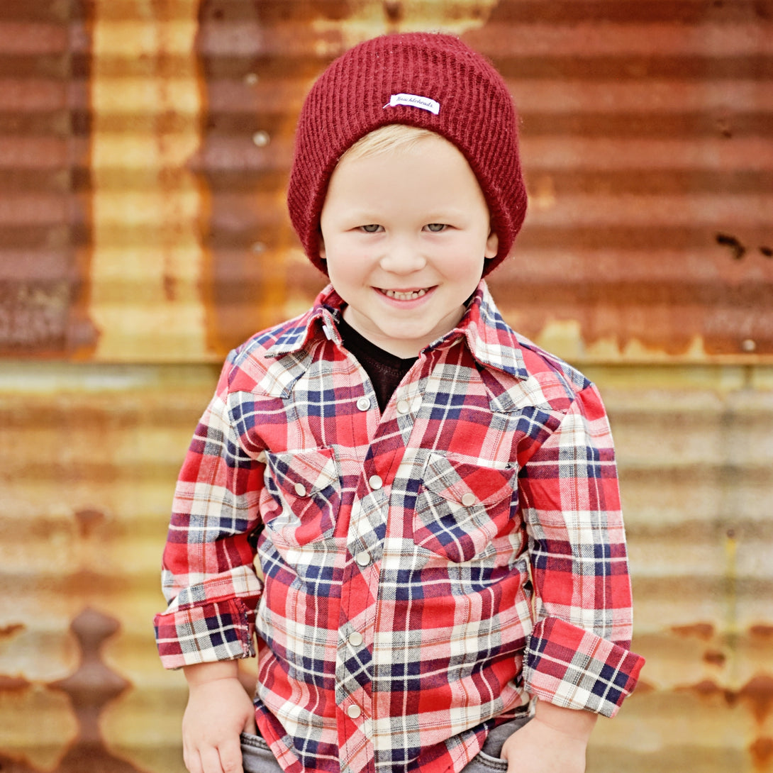 Image of a young boy confidently wearing a Knuckleheads Children's Burgundy Beanie, showcasing its adaptable and cozy design for infants and toddlers. Classic style with the Knuckleheads brand tag. This Toddler beanie is part of a collection of charming Infant hats.