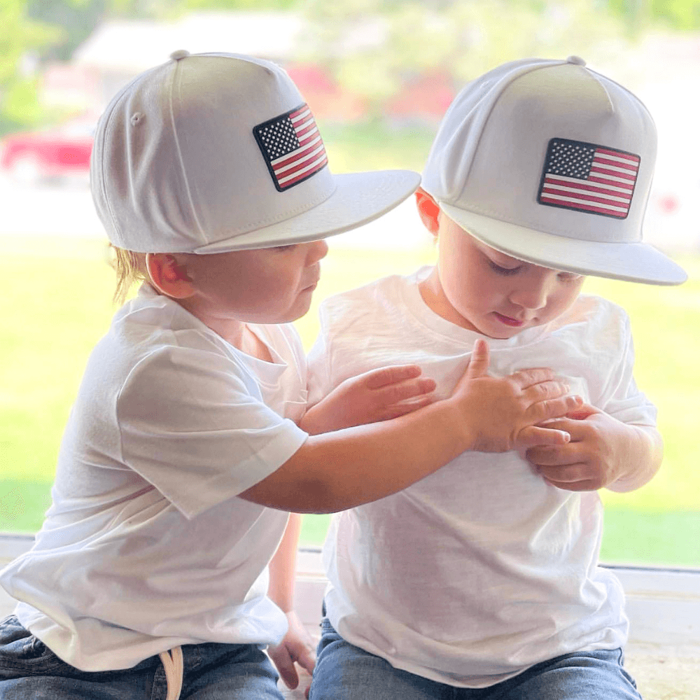 Image of White Trucker Hat for Kids with USA Flag Patch: A patriotic and stylish accessory designed for kids. In classic white, it showcases a prominent USA flag patch on the front. Elevate your child's style with this fashionable hat, perfect for showing their national pride. Crafted with care, this white kids trucker hat with the USA flag patch is a must-have addition to their wardrobe, suitable for various occasions and everyday wear.