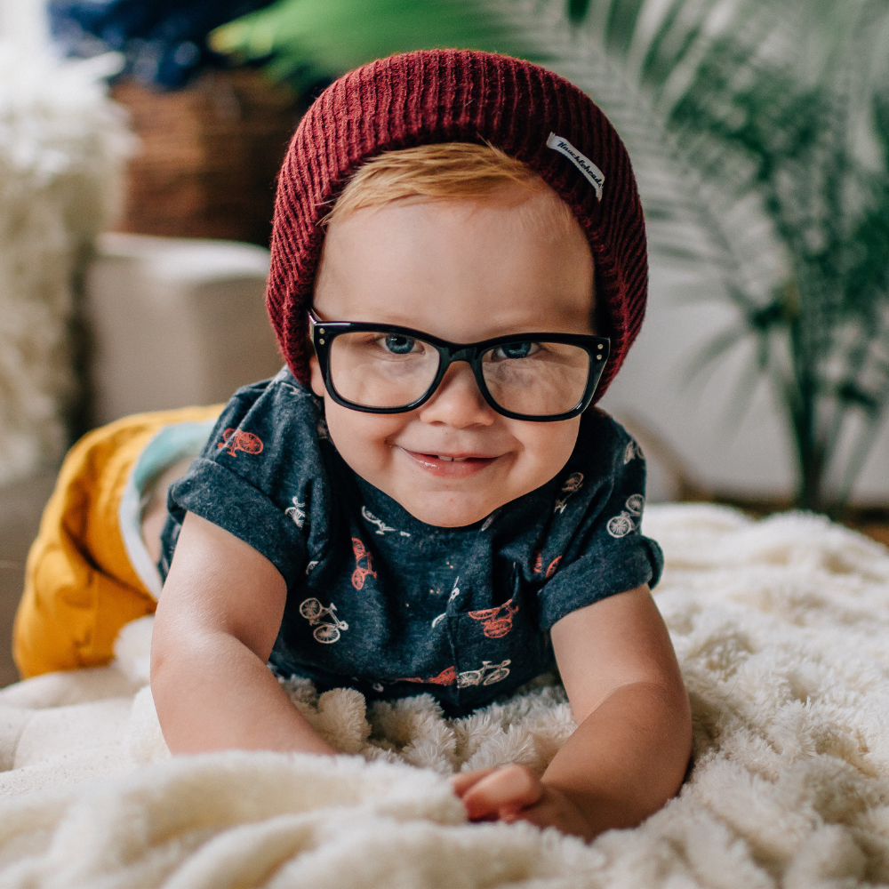 Image of a young boy confidently wearing a Knuckleheads Children's Burgundy Beanie, showcasing its adaptable and cozy design for infants and toddlers. Classic style with the Knuckleheads brand tag. This Toddler beanie is part of a collection of charming Infant hats.