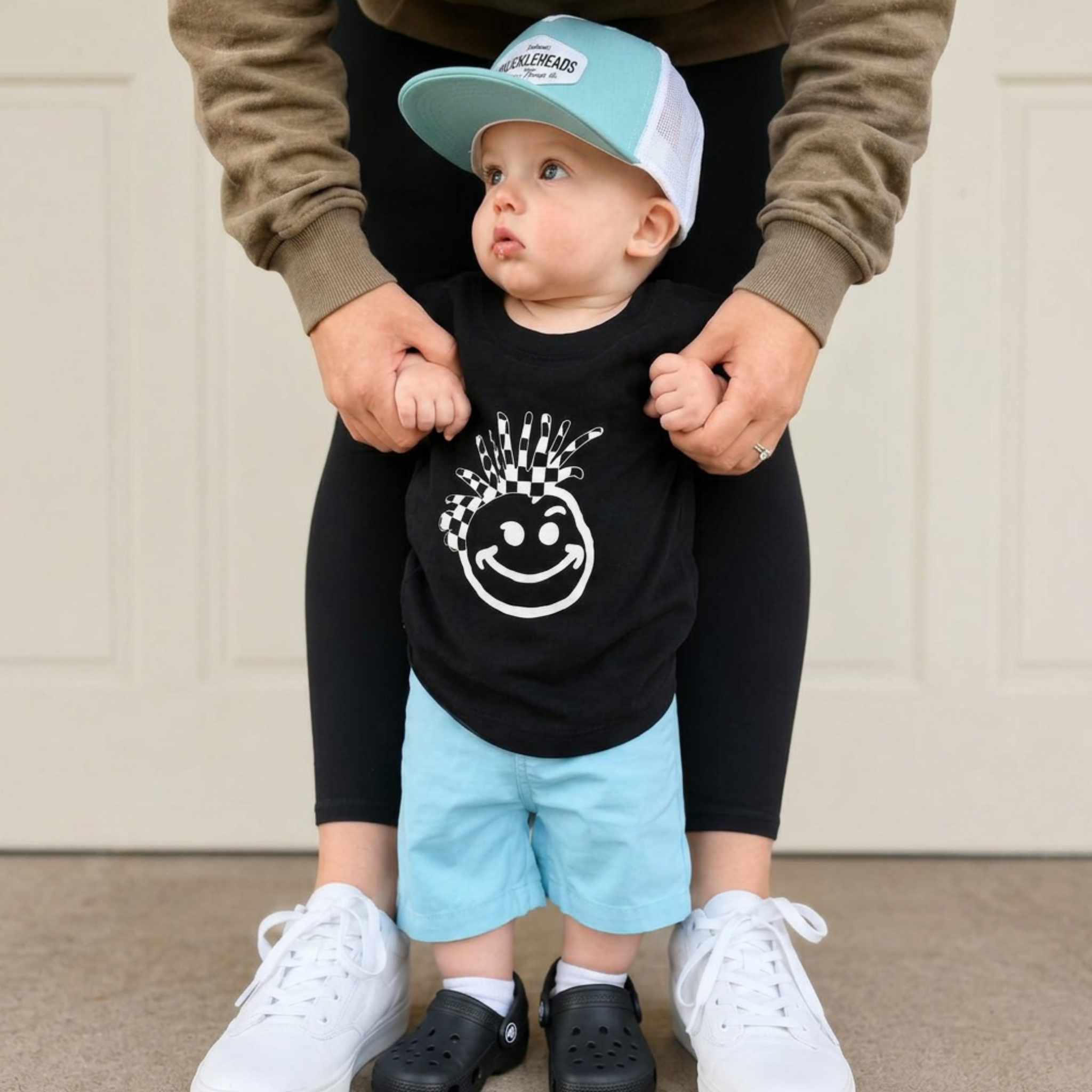 Toddler wearing FINAL SALE - Blue Knuckleheads Baby Kids Chino Shorts with a black graphic T-shirt and blue trucker hat while standing with support outdoors.