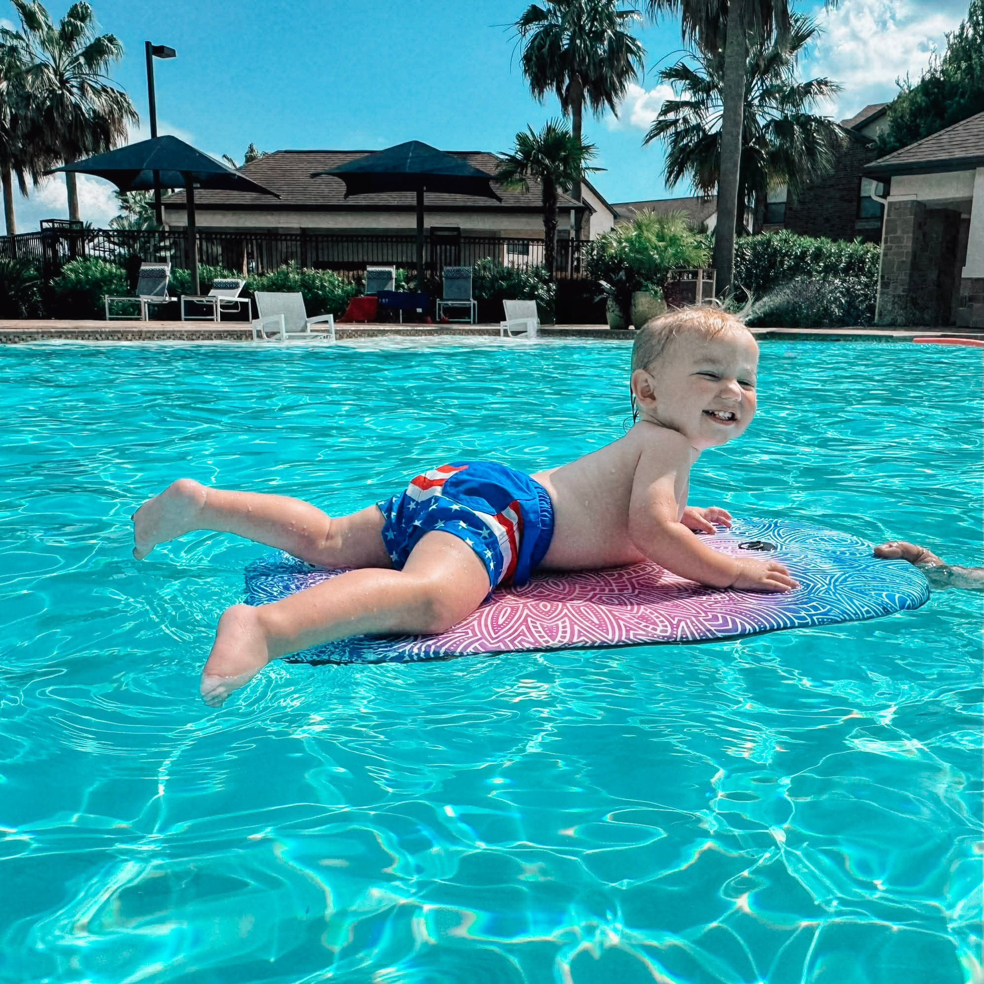 Toddler boy wearing Knuckleheads Retro USA flag swim shorts while floating in a swimming pool on a sunny summer day.