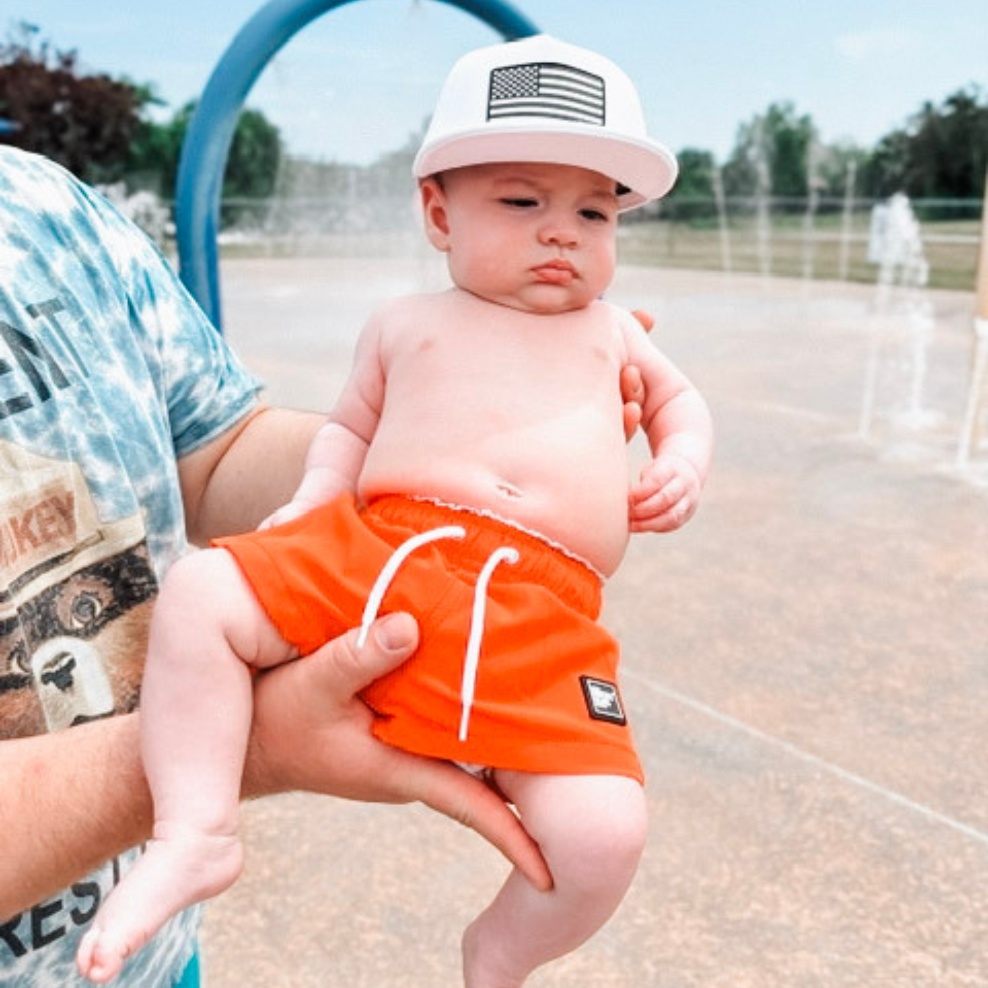 Toddler wearing bright orange swim shorts and a white American flag trucker hat at a splash pad, showcasing high-visibility kids swim trunks for summer water play.