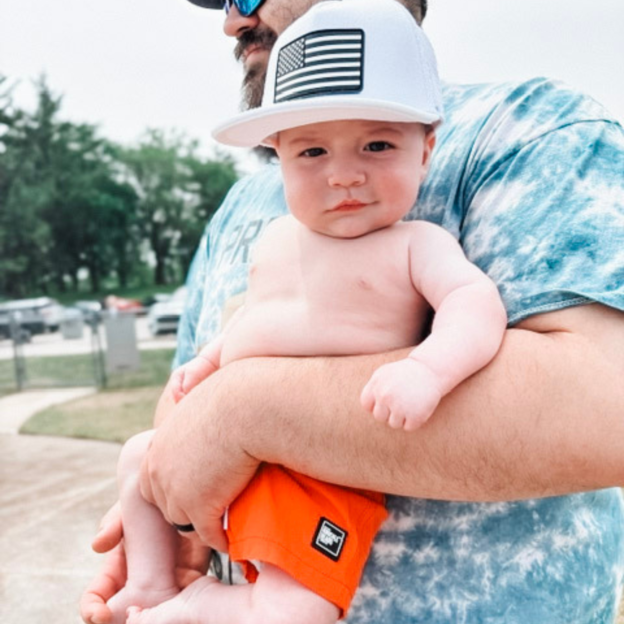 Baby wearing bright orange toddler swim trunks and a white trucker hat while being held outdoors, highlighting quick-dry, high-visibility swim shorts for kids.
