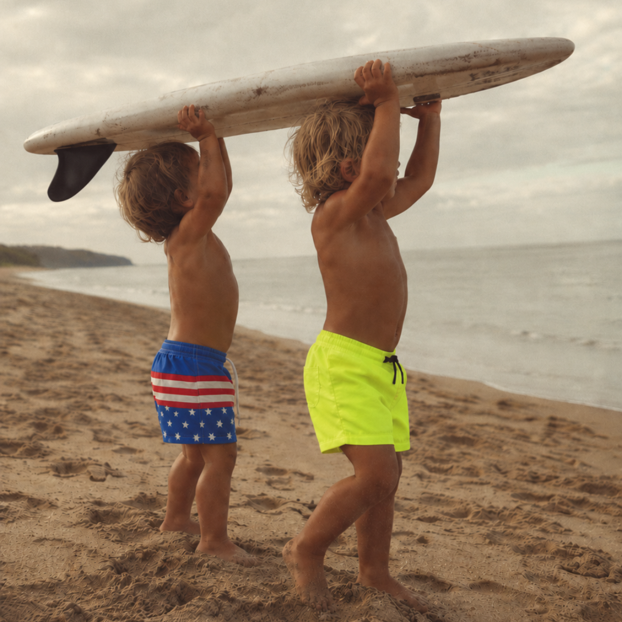 Child wearing Neon Yellow Swim Shorts while carrying a surfboard on the beach, with another child in Retro Swim Shorts beside them.