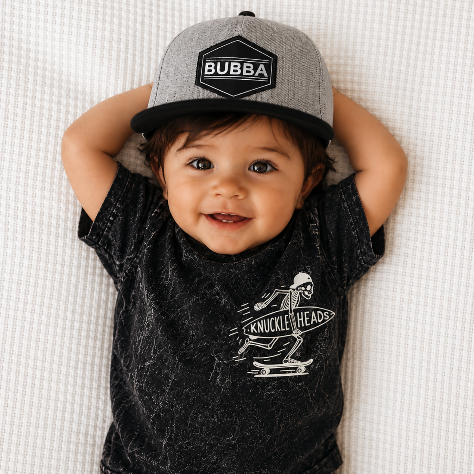 Toddler wearing the Bubba Lucas Trucker Hat with a gray front panel, black flat bill, and Bubba patch while smiling in a black graphic T-shirt.