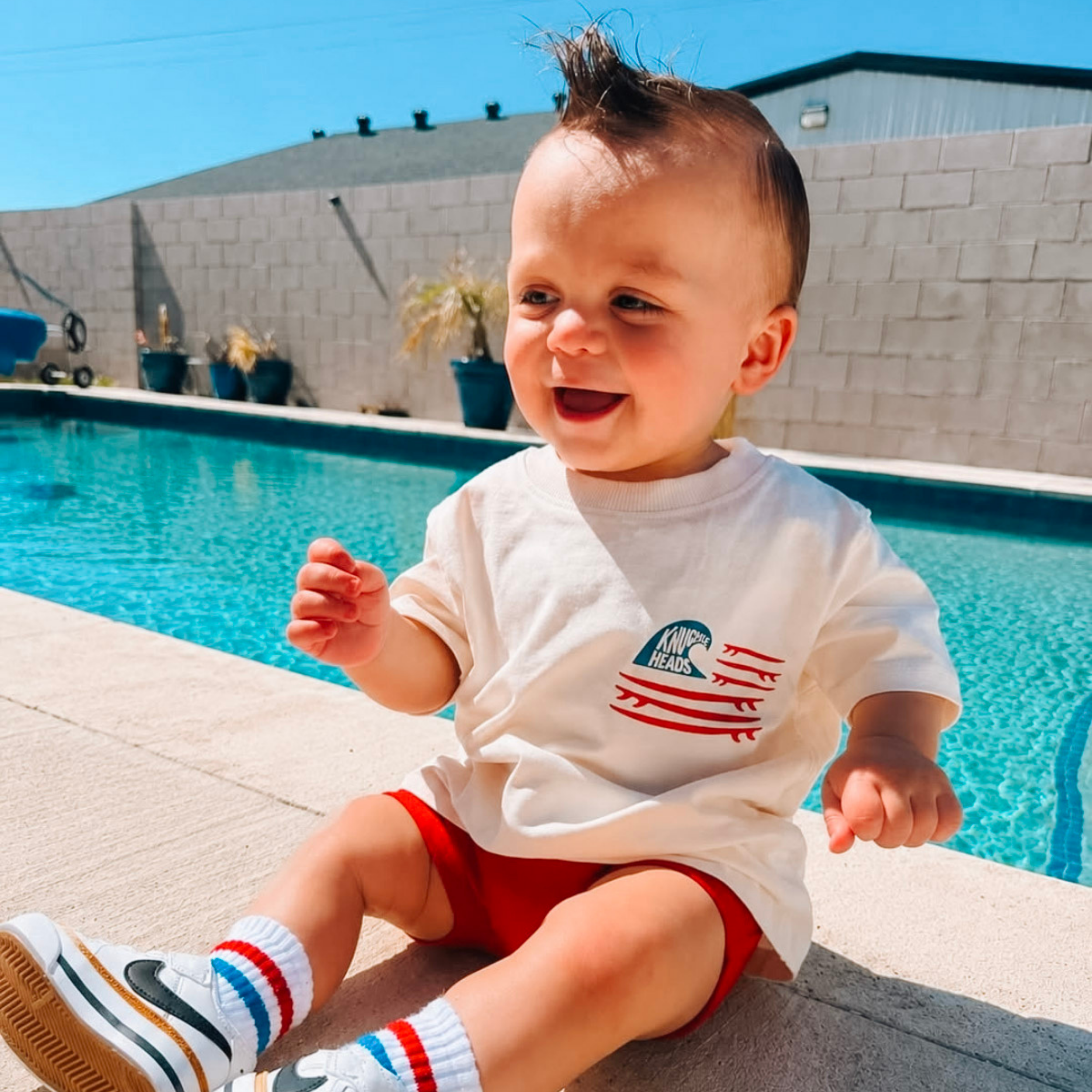 Toddler sitting poolside on a sunny day wearing the Knuckleheads Wave Rider Graphic Tee for Toddlers & Kids, red shorts, striped socks, and sneakers, smiling beside a swimming pool.