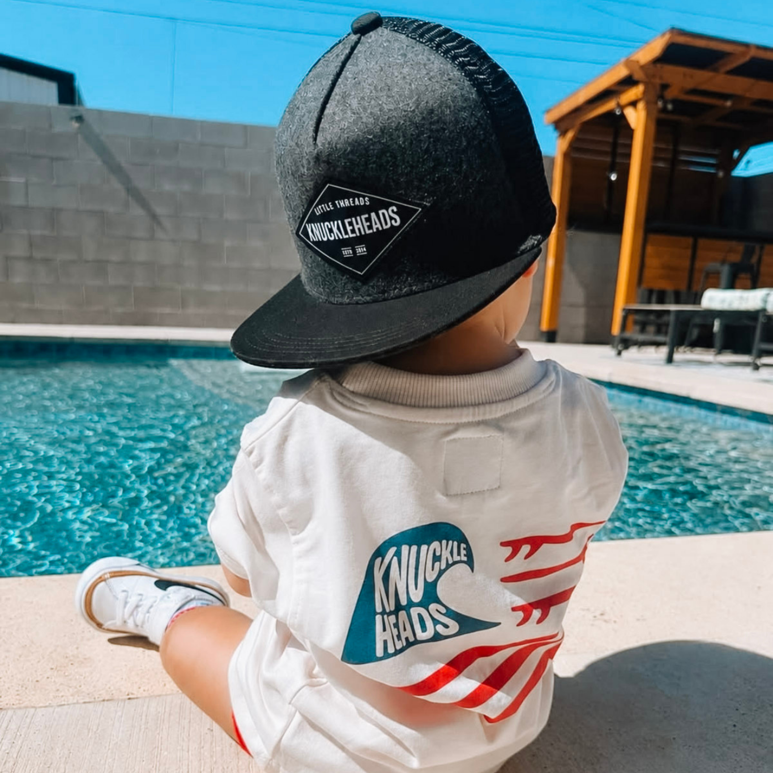 Back view of a toddler sitting by a swimming pool wearing a black Knuckleheads trucker hat and the Knuckleheads Wave Rider Graphic Tee for Toddlers & Kids, showcasing the Americana surf-inspired back graphic.