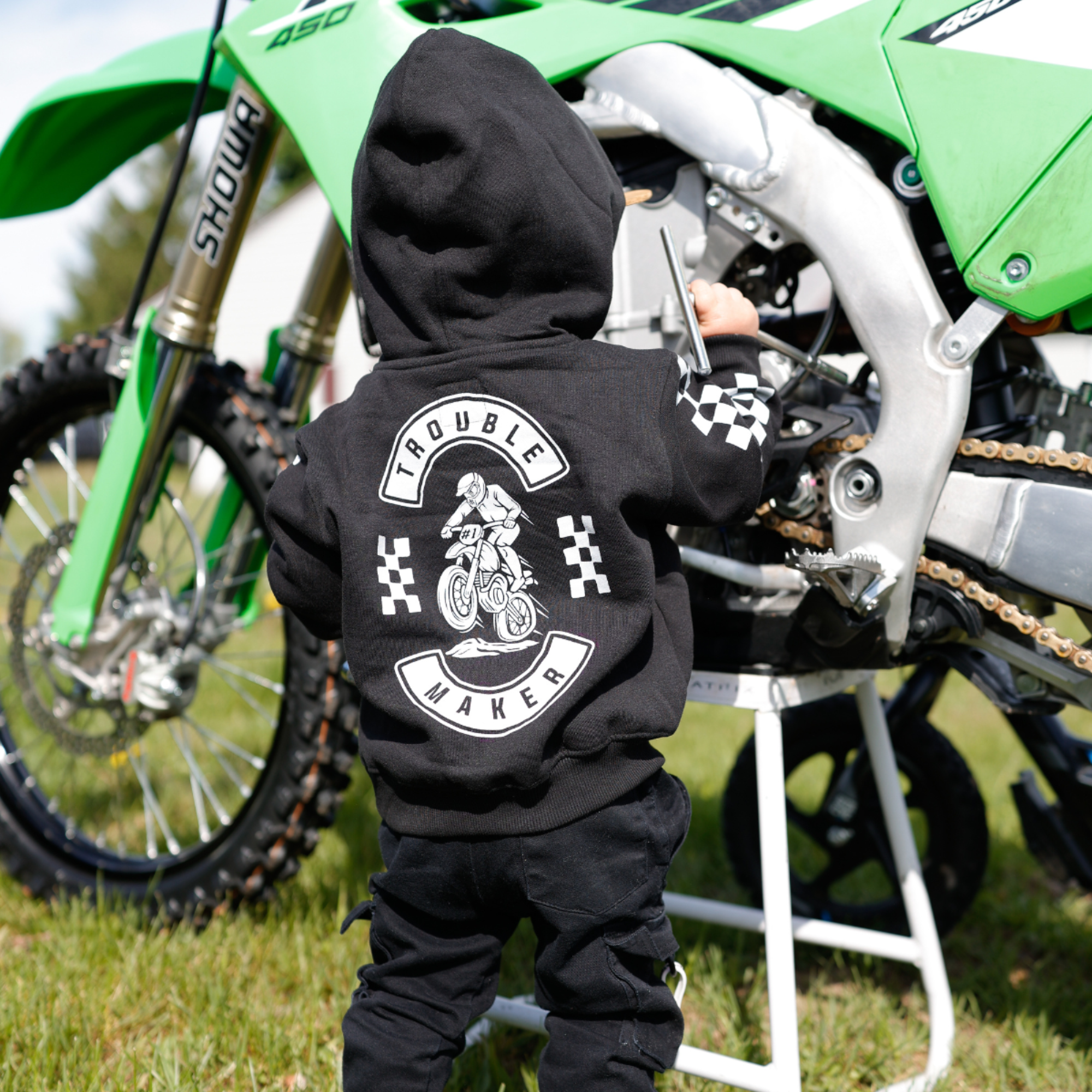 Toddler wearing a black Knuckleheads Trouble Maker motocross hoodie with large dirt bike graphic on the back while standing next to a motocross bike.