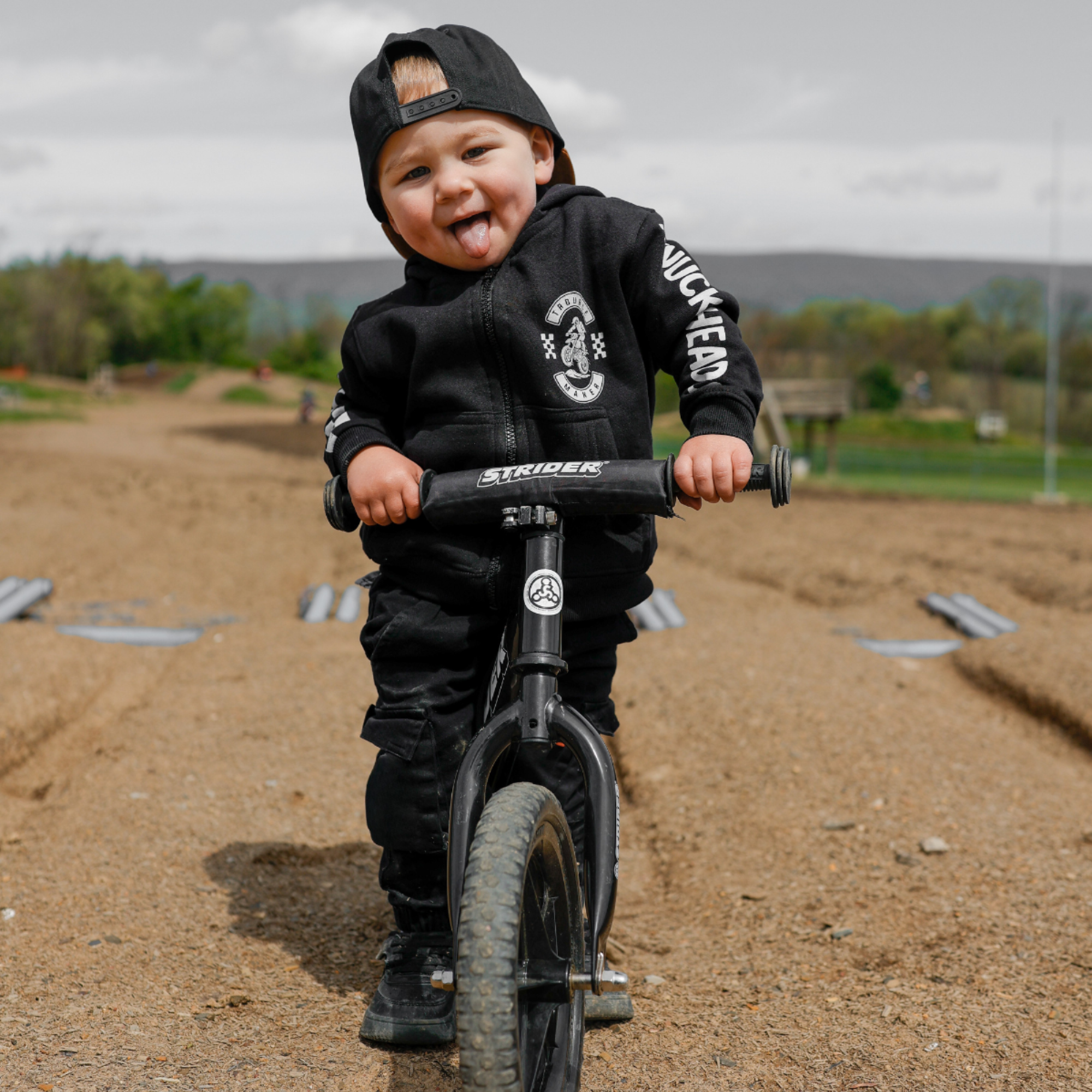Toddler wearing a black Knuckleheads Trouble Maker motocross zip hoodie and trucker hat while riding a balance bike outdoors.