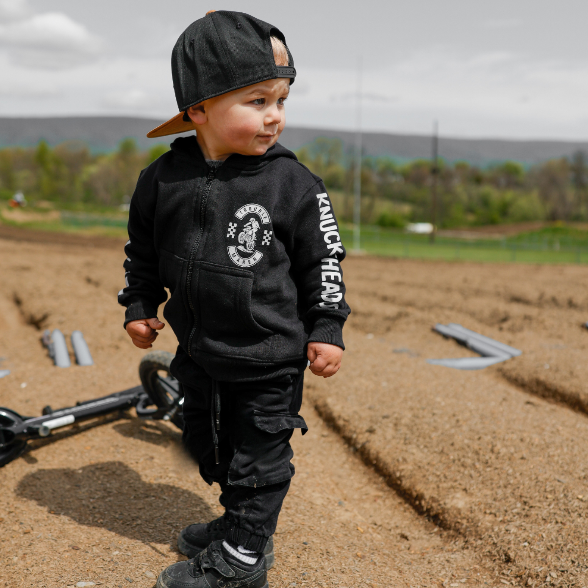 Toddler wearing a black Knuckleheads Trouble Maker motocross zip hoodie with racing graphics and checkerboard sleeve details outdoors at a dirt bike track.
