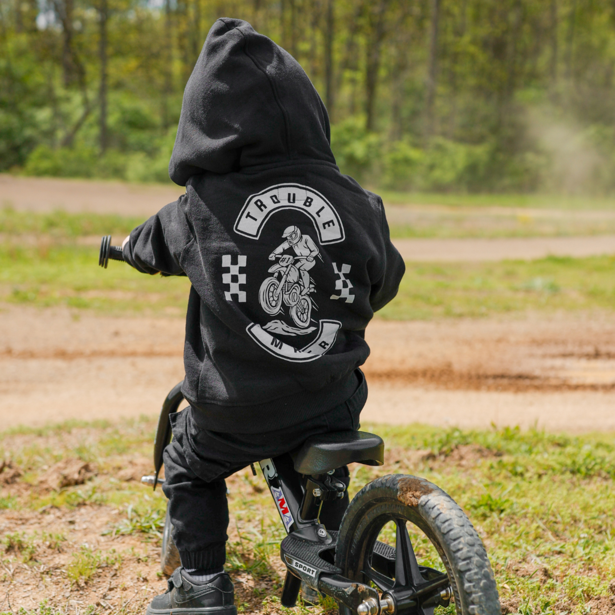Toddler wearing a black Knuckleheads Trouble Maker motocross zip hoodie with racing graphics and checkerboard sleeve details outdoors at a dirt bike track.