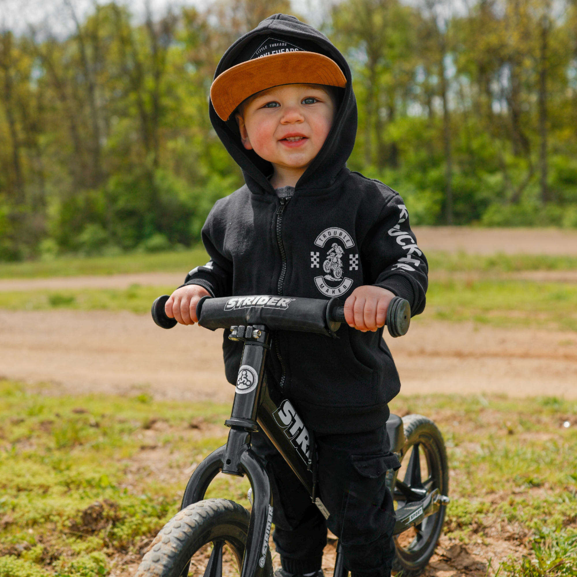Toddler wearing a black Knuckleheads Trouble Maker motocross zip hoodie with racing graphics and checkerboard sleeve details outdoors at a dirt bike track.