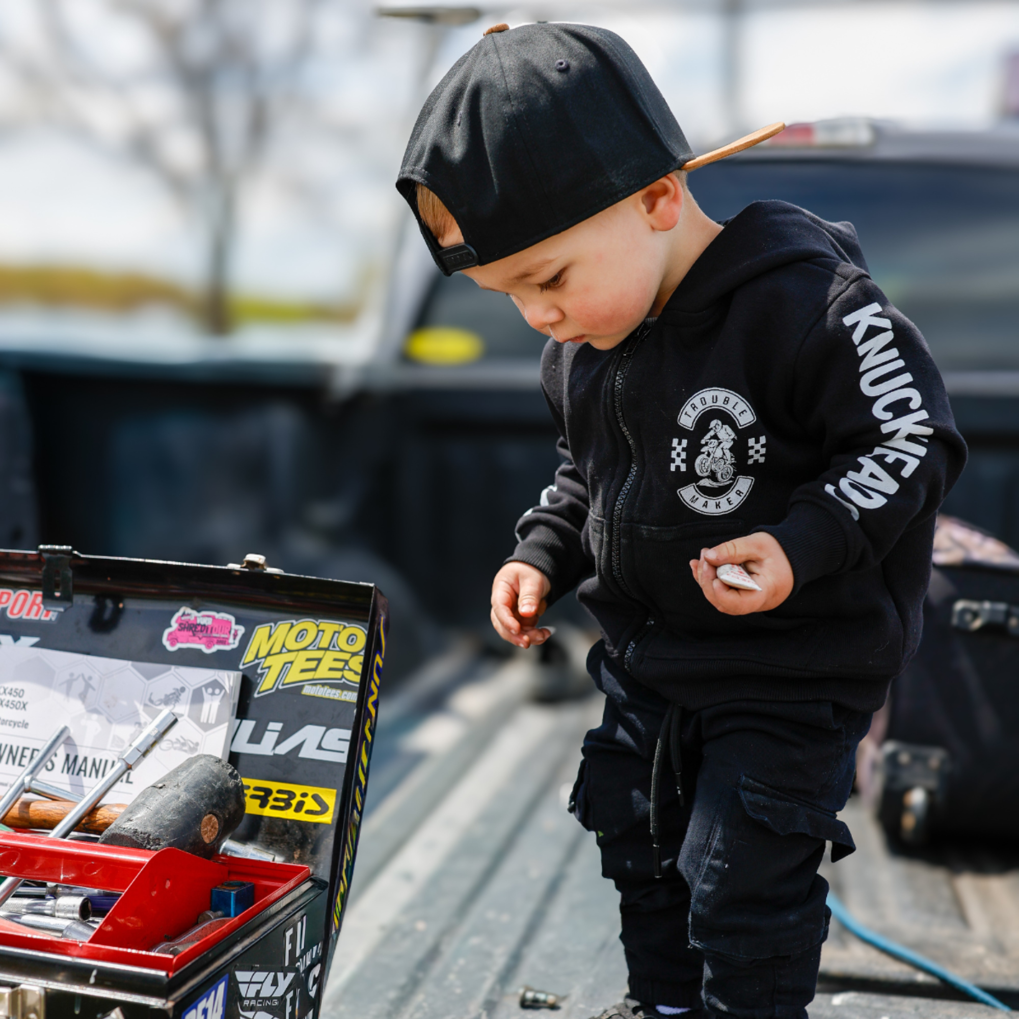 Toddler wearing a black Knuckleheads Trouble Maker motocross hoodie and snapback hat while standing next to a toolbox outdoors.