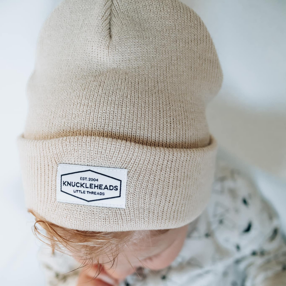 Image of Tan Kids Beanie with Knuckleheads Logo: This green beanie is both stylish and cozy for kids. The Knuckleheads logo on the front adds a unique touch to their outfits while keeping them warm. Crafted with care, this beanie is a versatile addition to their winter wardrobe, suitable for various occasions and everyday wear.