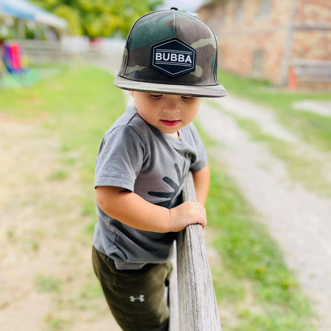 Presenting the Adventurous Kids Trucker Hat with 'Bubba' Patch: A trendy and stylish accessory designed for kids. In a cool camo pattern, it features a fun 'Bubba' patch on the front. Elevate your child's style with this fashionable hat, perfect for adding a touch of outdoor charm to their outfits.