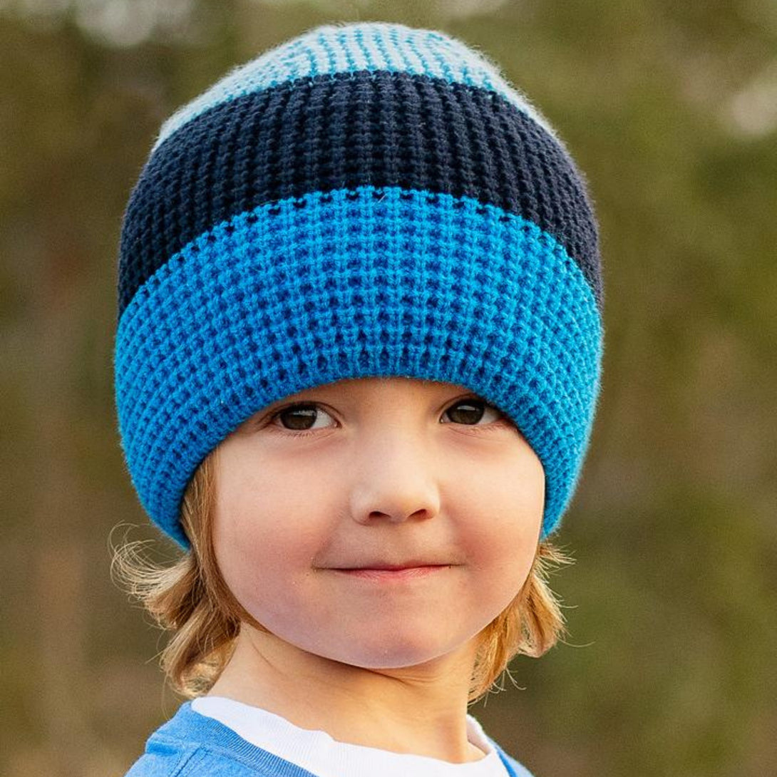 Image of a young boy confidently sporting a Knuckleheads Children's Blue Beanie, highlighting its adaptable and cozy design for infants and toddlers. Classic style featuring the Knuckleheads brand tag. This beanie for toddlers is part of a collection of delightful infant hats.