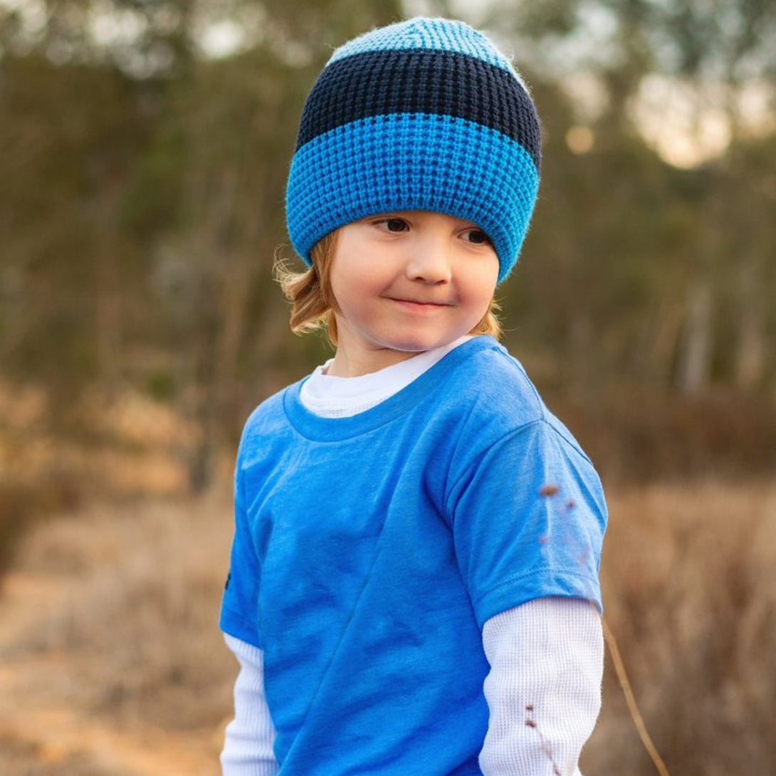 Image of a young boy confidently sporting a Knuckleheads Children's Blue Beanie, highlighting its adaptable and cozy design for infants and toddlers. Classic style featuring the Knuckleheads brand tag. This beanie for toddlers is part of a collection of delightful infant hats.