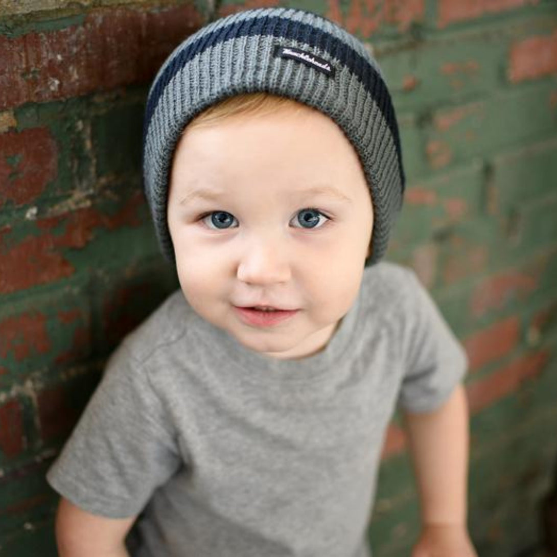 Young boy with a playful smile wearing a black and grey striped beanie adorned with a Knuckleheads tag, exuding a cool and youthful vibe.