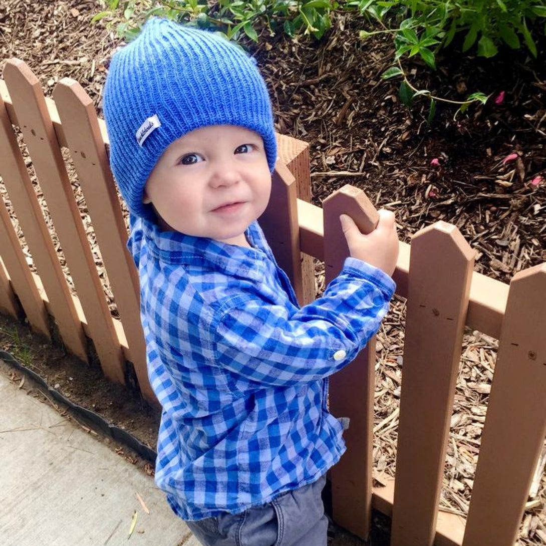 Image of a young boy wearing a children's blue Baby Beanie by Knuckleheads, showcasing its versatile and comfortable design for infants and toddlers. Classic style with Knuckleheads brand tag. This Toddler beanie is part of a collection of charming Infant hats.