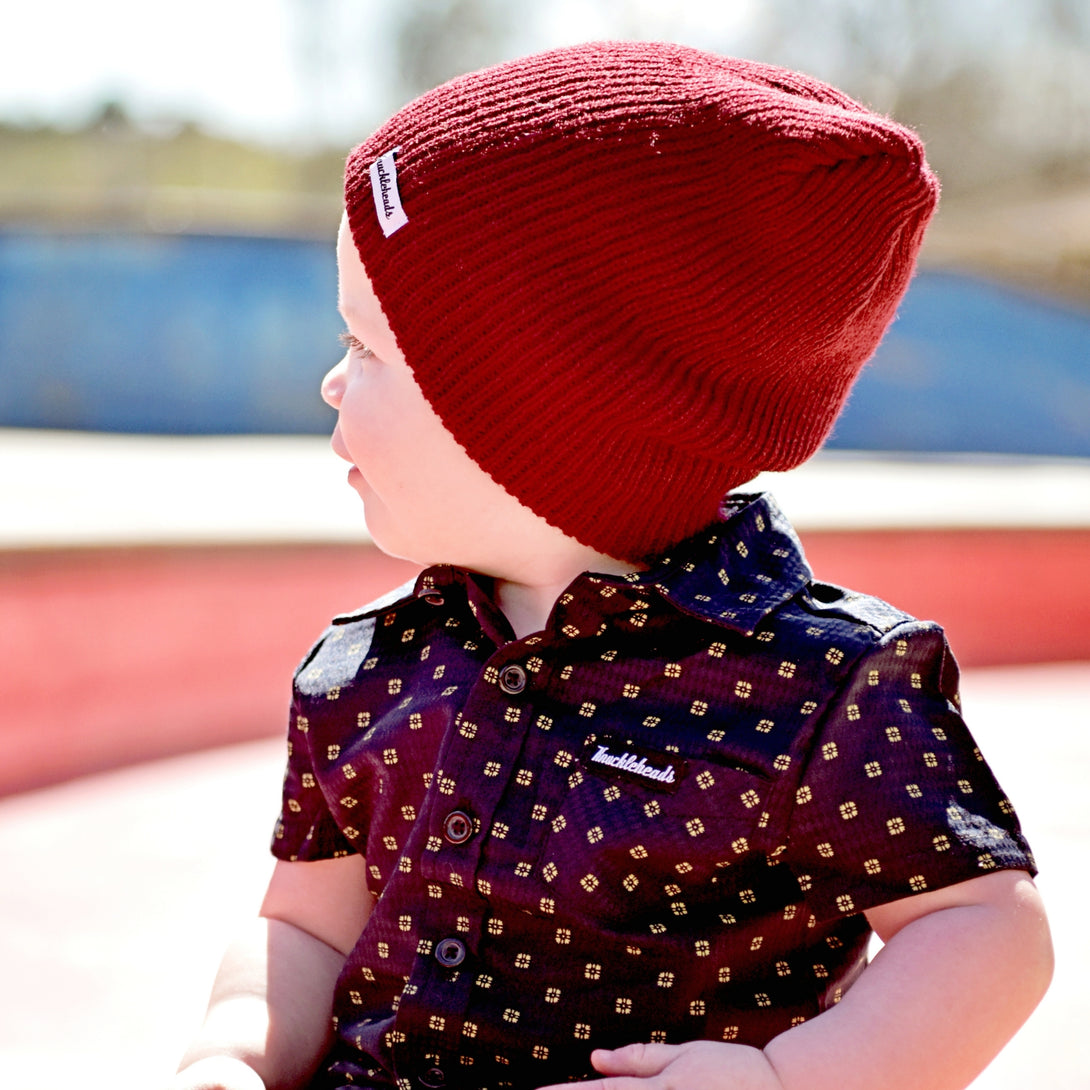 Image of a young boy confidently wearing a Knuckleheads Children's Burgundy Beanie, showcasing its adaptable and cozy design for infants and toddlers. Classic style with the Knuckleheads brand tag. This Toddler beanie is part of a collection of charming Infant hats.