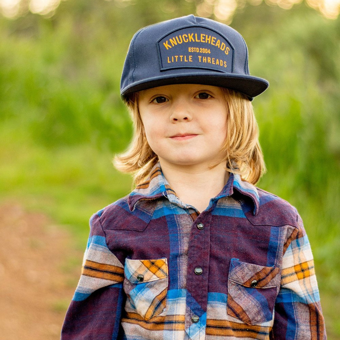 Image of Navy Kids Trucker Hat with Knuckleheads Patch: A stylish and versatile accessory designed for kids. In deep navy, it showcases a striking Knuckleheads patch on the front. Elevate your child's style with this fashionable hat, perfect for adding a touch of character to their outfits. Crafted with care, this navy kids trucker hat with the Knuckleheads patch is a must-have addition to their wardrobe, suitable for various occasions and everyday wear.
