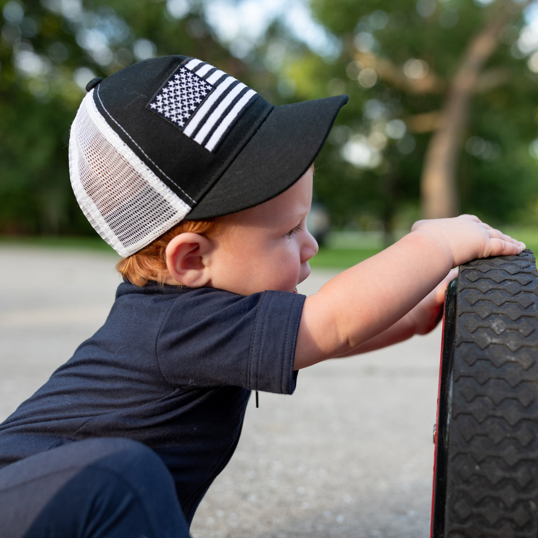 Image of Black Kids Trucker Hat with White Mesh and USA Flag Patch: A patriotic and stylish accessory designed for kids. In deep navy with crisp white mesh, it features a prominent USA flag patch on the front. Elevate your child's style with this fashionable hat, perfect for adding a touch of national pride to their outfits while ensuring breathability.