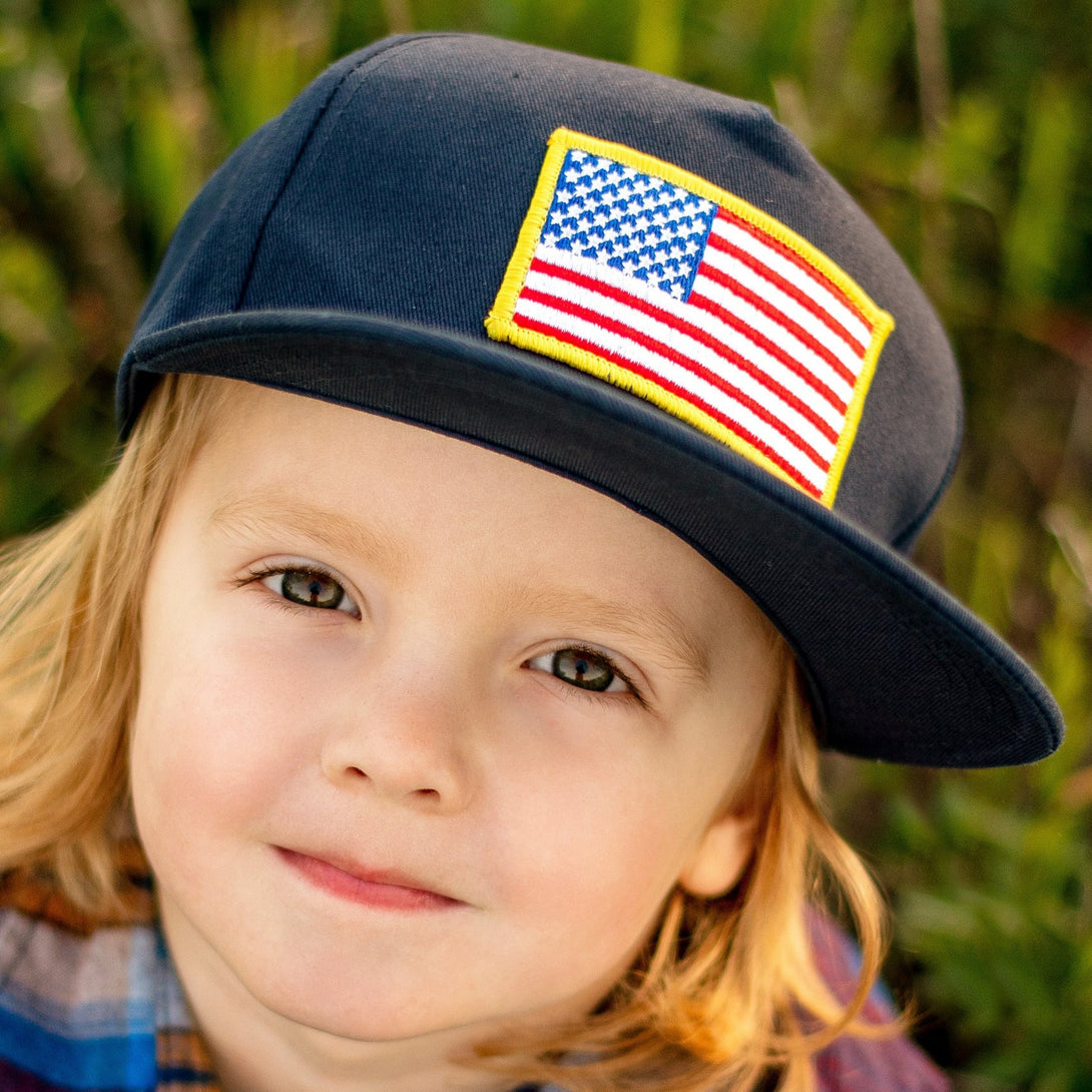 Image of Navy Kids Trucker Hat with USA Flag Patch: A patriotic and stylish accessory designed for kids. In deep navy, it features a prominent USA flag patch on the front. Elevate your child's style with this fashionable hat, perfect for adding a touch of national pride to their outfits. 